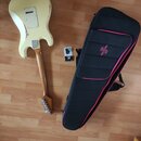 Kremově bílá elektrická kytara s tvarem typu Stratocaster, položená na dřevěné podlaze. Na zadní straně těla jsou viditelné oděrky a opotřebení laku.…