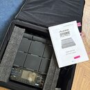 Černý sampling pad značky ALESIS v přepravním kufru s polstrováním. Vedle nástroje leží uživatelský manuál s nápisem ALESIS STRIKE MULTIPAD SAMPLING…