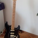 Černá elektrická kytara s bílým pickguardem a javorovým hředníkem. Na hlavě je viditelné logo Fender a nápis STRAT. Nástroj vykazuje běžné známky pou…
