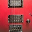 Detail červené elektrické kytary s dvěma humbuckery a strunní kotvou. Na těle je v horní části vidět část kulatého otvoru s ozdobnou vložkou. Povrch…