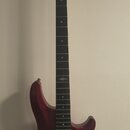 Elektrická basová kytara s červeným kovovými lakem a černým hmatníkem. Na hlavě je bílé logo Sgr. Tělo nástroje obsahuje dva humbuckery, čtyři ovláda…