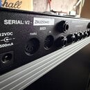 Detail zadní strany černého elektronického zařízení s řadou konektorů. Je zde vidět nápis SERIAL: V2 a nálepka s číslem ZMJ250443, dále označení MIDI…