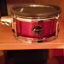 Elektronický snare s fialovým třpytivým povrchem a chromovanými obručemi. Na přední straně je viditelné logo ALESIS v kruhovém medallionu a stejný ná…