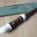 Hnědá sopránová bloková flétna s bílými plastovými spojkami a mouthpiecem. Na těle nástroje je vyryto logo YAMAHA. Vedle flétny je zelený textilní po…