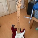 Kytara s červeným bodies a světlým dřevěným hmatníkem. Na hlavě je viditelné logo "Fender". Instrument má bílý pickguard a tři snímače. Kytara stojí…