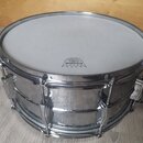 Stříbrný kovový snare drum s bílým napnutým blanou. Na bláně je černý nápis WEATHERKING AMBASSADOR COATED. Povrch korpusu vykazuje známky opotřebení…