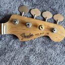 Hlava basové kytary z přírodního dřeva s čtyřmi kovovými ladičkami. Na dřevě je černý nápis Fender a text JAZZ BASS ELECTRIC BASS. Povrch dřeva vykaz…