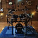 Elektronická bicí souprava značky Roland s černými bubny a činely. Na basovém bubnu je bílý nápis Roland a V-Drums. Souprava je umístěna na černém ko…