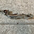 Chromovaný kovový mechanismus kytarového tremola. Na povrchu je viditelné jemné opotřebení a drobné škrábance. Vedle nástroje leží stříbrný kovový ps…