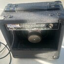Zadní strana kytarového komba v černém texturovaném povrchu s rukojetí. Na zadní straně je bílá nálepka s nápisem CASH GENERATOR a varovným textem, p…