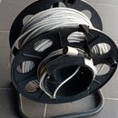 Černý plastový náviják s kovovým spodním rámem, na kterém je namotán šedý kabel. Na kabelu je viditelný konektor. Nástroj nemá žádné viditelné značky…