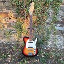 Elektrická kytara s tělem v provedení sunburst a bílým pickguardem. Na hlavě hředu je viditelné logo "Fender". Nástroj vykazuje známky opotřebení, ze…