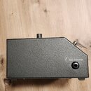 Černý kovový přístroj s texturovaným povrchem a asymetrickým tvarem. Na pravé straně je umístěn konektor s nápisem FROM AMPLIFIER. Horní plocha obsah…