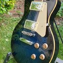 Tmavě modrá elektrická kytara typu Les Paul s krémovým pickguardem a dvěma humbuckery. Nástroj je umístěn na černém stojanu v travnatém terénu. Na ky…