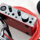 Červený audio rozhraní s nápisem Scarlett. Na horní straně jsou dva otočné knoflíky, jeden velký ovládací prvek a několik vstupů a tlačítek s nápisy…