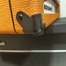 Detailní záběr na roh oranžového kytarového komba s texturovaným povrchem. Na boku je připevněn černý plastový držák na kabel. Pod kombem je vidět čá…