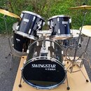 Černá bubnová sada s kovovým kováním a zlatými činely. Na basovém bubnu, tom-tamech i snare je viditelný nápis SWINGSTAR by TAMA. Na jednom z činelů…