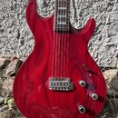 Elektrická kytara s červeným lakovaným tělem s viditelnou strukturou dřeva. Nástroj má dva humbuckery, tři ovládací knofíky a přepínač poloh snímovač…