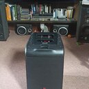 Černý reproduktor s mřížkou v přední části. Ve spodní části mřížky je umístěno červenobílé logo JBL. Horní strana přístroje obsahuje ovládací prvky a…