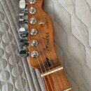 Detail hlavy a hředu elektrické kytary z dřeva s přirozeným povrchem. Na hlavě je vidět nápis "Fender" a modelové označení "TELECASTER". Nástroj má š…