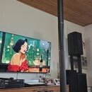 Černý reproduktorový systém složený z vysokého sloupového reproduktoru a subwooferu. Na přední straně subwooferu je bílý nápis JBL. V pozadí jsou vid…