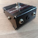 Černý kovový efektový pedál s nápisem SWITCH BOX na horní straně. Na vrchu je umístěn jeden kovový spínací tlačítko. Přední strana obsahuje tři konek…