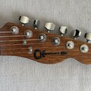 Detail hlavy kytary z hnědého dřeva s viditelnou strukturou slázy. Na hlavě je černý nápis „Charvel“ s doprovodným logem. Nástroj má šest chromovanýc…