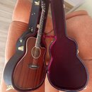 Akustická kytara s hnědým dřevěným povrchem v černém tvrdém kufru s burgundy vnitřkem. Na hlavě kytary je viditelné logo, které je však nečitelné. Ná…