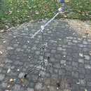Kovový stativ s trojnožkou a teleskopickým ramenem. Na konci ramene je umístěn malý modrý váleček s kovovým zakončením. Na stativu nejsou viditelné ž…