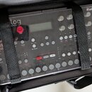 V černé přepravní tašce jsou upevněny dva elektronické přístroje s průhledným plastovým krytem. Levý přístroj nese nápis "analog Synthesizer" a má če…
