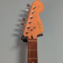 Fotografie zachycuje hlavu a hриф elektrické kytary v přirozené dřevěné barvě. Na hlavě je vidět nápis Squier by Fender a STRAT. Povrch dřeva vykazuj…