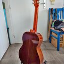 Zadní pohled na akustickou kytaru s lakovaným povrchem v červenohnatém odstínu. Nástroj je umístěn na černém stojanu. Na fotce nejsou žádné viditelné…