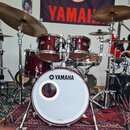 Bicí souprava v tmavě červené barvě s kovovým hardwarem. Na bílém předním z koženě basového bubnu je čitelné logo a nápis YAMAHA. V pozadí visí černý…