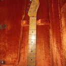 Kytara s hřednou deskou z jasného dřeva v oranžovém pouzdře. Na hlavě je viditelné logo Fender a nápis STRATOCASTER. Hриф má tmavé tečkové značky pol…