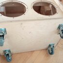 Světle hnědý dřevěný box z překližky s dvěma velkými kruhovými otvory v horní části. Na spodní straně jsou připevněna čtyři kovová kolečka s šedými p…