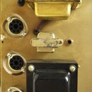 Detail vnitřku elektronického zařízení na zlatavém kovovém šasi. Jsou zde vidět tři kulaté vícepinové konektory, dva transformátory a jeden menší kov…