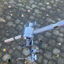 Detail chromovaného spojovacího mechanismu stativu s dvěma knírkovými šrouby. Z mechanismu vychází jedna kovová tyč a druhá tyč potažená šedou textur…
