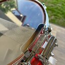 Detailní záběr na červený buben s kovovým obručem a mechanismusem snare throw. Na kovovém prvku je viditelné logo Pearl. Povrch kovu je chromovaný a…