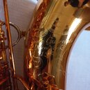 Detail zlatavého, lesklého povrchu saxofonu s rytým ornamentem. Na těle nástroje je vyryt nápis „King Guardala New York USA“. Povrch je v dobrém stav…