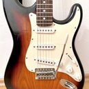 Elektrická kytara s tělem v provedení sunburst a bílým pickguardem. Nástroj má tři snímače s bílými kryty, chromovaný most s tremolem a bílé ovládací…