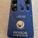 Modrý kytarový efekt s nápisy JOYO a ARGOS OVERDRIVE. Na horní straně jsou tři velké černé otočné knoflíky s označením ATTACK a LEVEL, dvě menší čern…