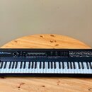 Černý digitální syntetizátor s klaviaturou, umístěný na dřevěném stole. Na levé straně je bílý nápis Roland D-50 Linear Synthesizer. Horní panel obsa…