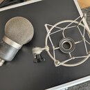 Na fotografii je stříbrný mikrofon s kovovou síťkovou hlavou a doprovodný šokabsorber (pavouk) v odpovídající stříbrné barvě. Povrch těla mikrofonu v…