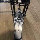 Fotografie zachycuje detail bubnového pedálu z kovového materiálu s kartáčovaným povrchem. Na základní desce je umístěno oválné logo s nápisem MAPEX.…