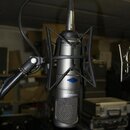 Stříbrný kondenzátorový mikrofon umístěný v černém pavouku. Na těle mikrofonu je svislý nápis TAKSTAR CM-450 a modrý kosočtvercový symbol. Povrch mik…