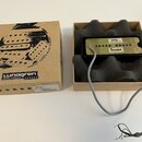 Kartonová krabice s nápisem Lundgren guitar pickups a grafickým motivem. Vedle ní je v krabici s pěnovou výplní černý kytarový snímač se zlatým kryte…