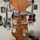 Záběr na zadní stranu dřevěné hlavy kytary s šesti chromovanými ladičkami. Na každém z ladicích mechanismů je vyryto logo Ovation. Povrch dřeva je la…