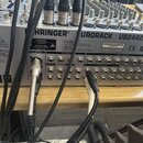 Zadní strana stříbrného mixážního pultu s nápisy BEHRINGER a EURORACK UB2442FX-PRO. Jsou vidět různé typy konektorů, včetně XLR a jacků, do kterých j…