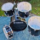 Černá bicí souprava složená z několika bubnů, položená na modré přikrývce. Na přední straně největšího bubnu je bílý nápis YAMAHA s doprovodným logem…
