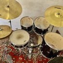 Sada bubnů v černé barvě s kovovými obručmi a zlatavými činely. Na bubnech jsou viditelné loga ZAMA, na jednom z činelů je nápis PAISTE 302 a na jiné…